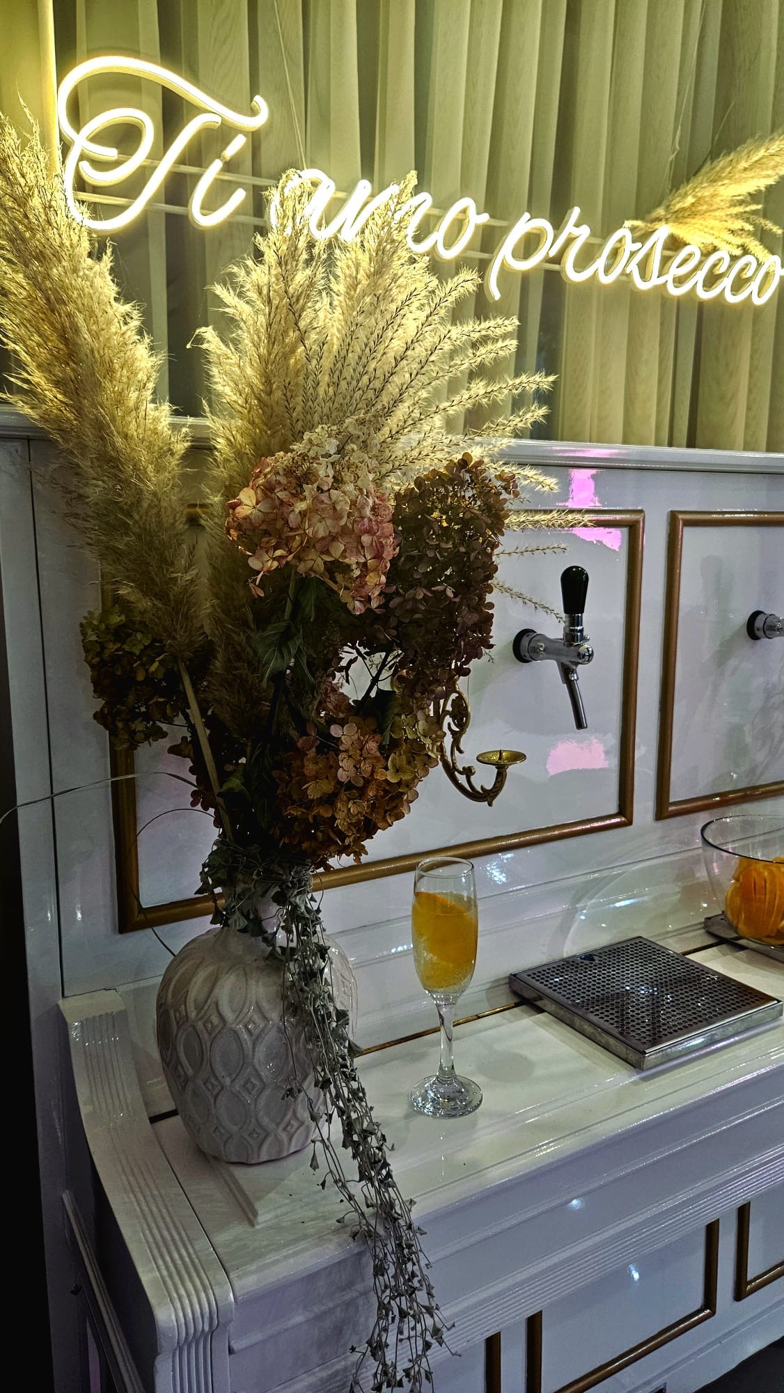 Dekoracja baru z prosecco: wysoki wazon z suszonymi trawami i hortensjami, neonowy napis 'Ti amo prosecco', szklanka prosecco z pomarańczą, biały bar z kranami do nalewania.