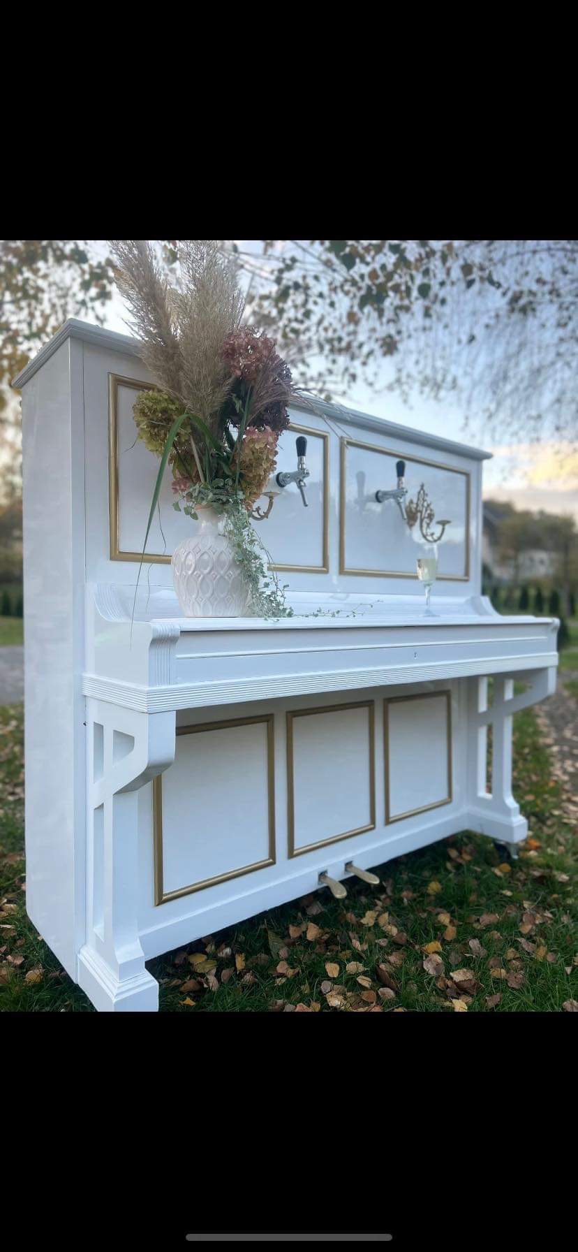 Białe pianino przerobione na bar z kranami do piwa, ozdobione złotymi ramkami i wazonem z suszonymi kwiatami, stojące na trawie.