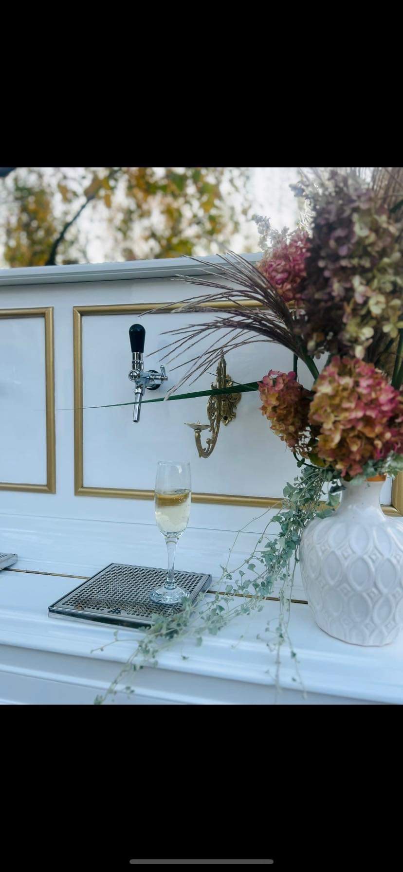 Elegancki bar z białym frontem, złotymi obramowaniami, kranem do piwa/napojów, szklanką z napojem, kratką ociekową i wazonem z suszonymi kwiatami, tworzący stylową aranżację.