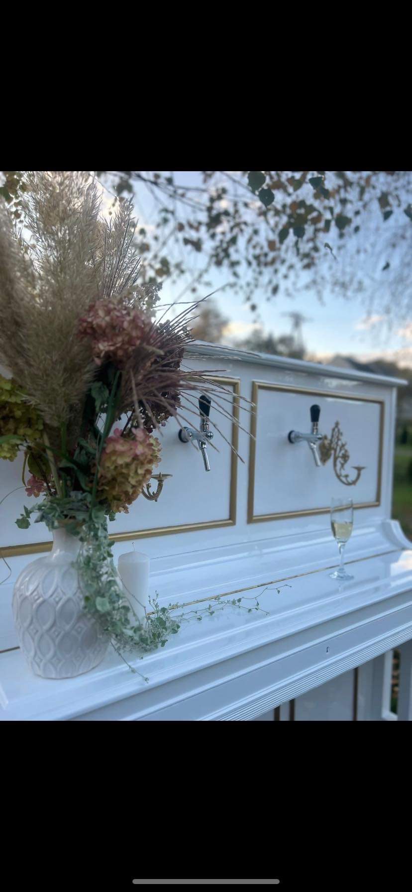 Biały pianino-bar z dwoma kranami, dekoracją kwiatową w wazonie, świecą i kieliszkiem szampana na tle plenerowym.