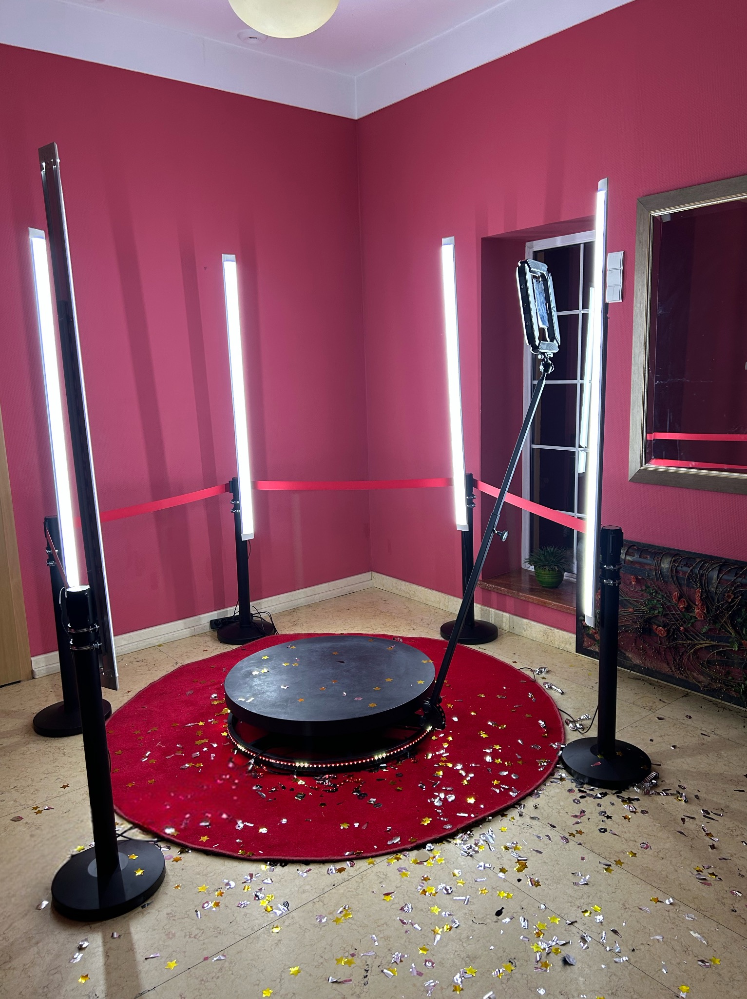 Oświetlona platforma obrotowa na czerwonym dywanie otoczona czterema lampami na statywach i konfetti w pomieszczeniu z bordowymi ścianami, prawdopodobnie gotowa do sesji zdjęciowej lub nagrywania...
