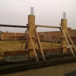 Wznoszenie fundamentów i ścian z cegły silikatowej pod budynek, widoczne drewniane szalunki pod betonowe słupy oraz zbrojenie.
