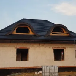 Niedokończony dom z białego pustaka z ciemnym dachem z dwoma półokrągłymi oknami dachowymi w trakcie montażu; widoczne rusztowanie i zbiornik na wodę.