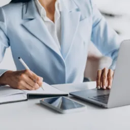 Kobieta w błękitnym żakiecie notuje w notatniku obok laptopa i smartfona na białym biurku.