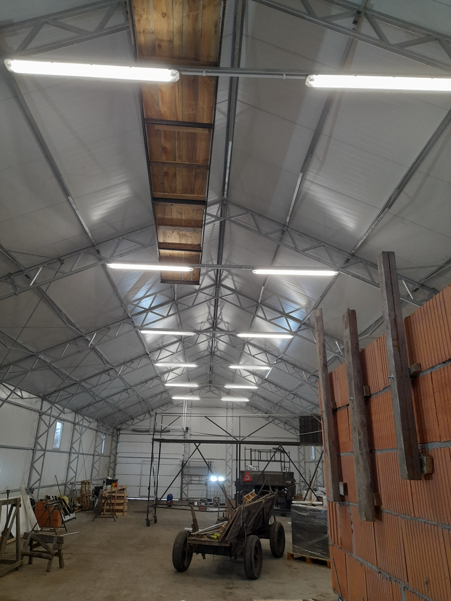 Wnętrze hali z metalową konstrukcją dachu, oświetleniem jarzeniowym i częściowo wymurowaną ścianą z cegły, widoczny wózek transportowy i rusztowanie.