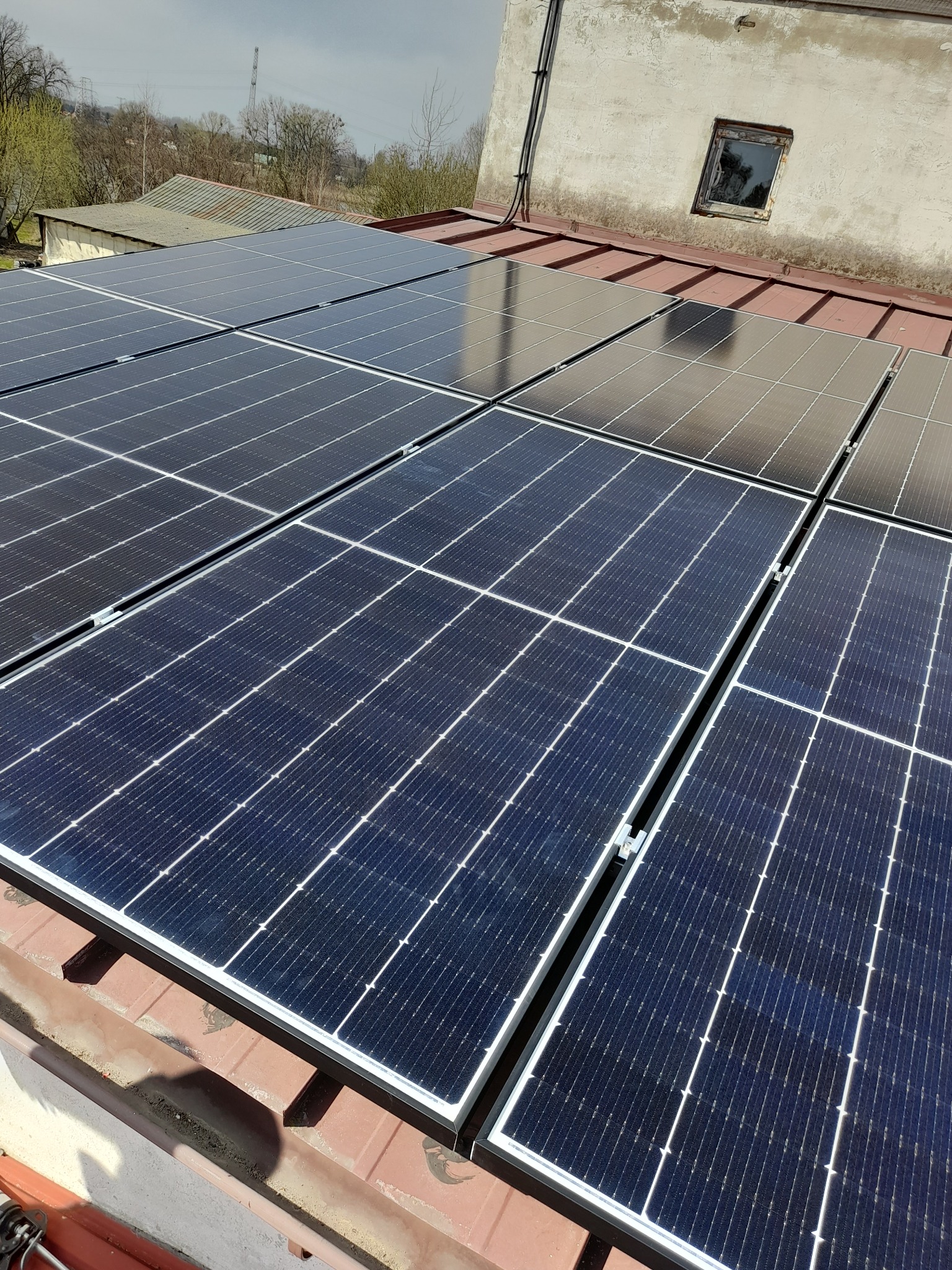 Zainstalowane panele fotowoltaiczne na czerwonym dachu budynku z widocznym starym oknem na ścianie.