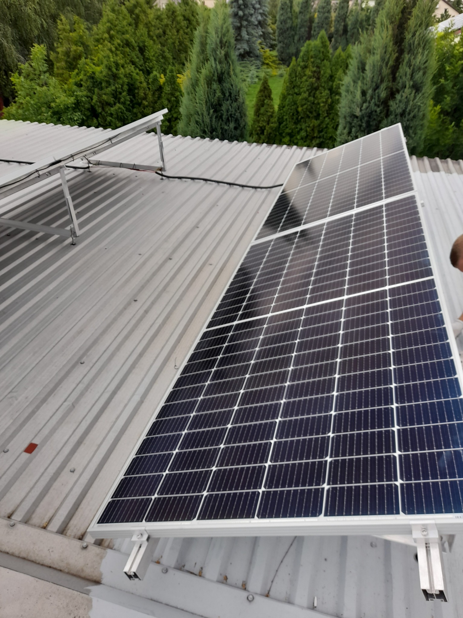 Panele fotowoltaiczne zamontowane na metalowym dachu z widocznymi mocowaniami i zielenią w tle.