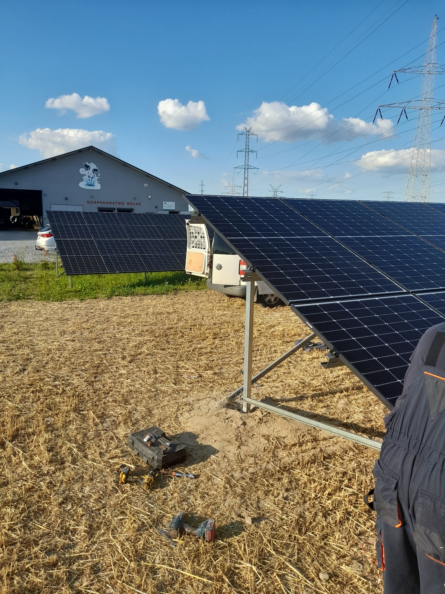 Montaż paneli słonecznych na polu, widoczny fragment kombinezonu roboczego, skrzynka z narzędziami i budynek gospodarczy z rysunkiem krowy w tle.