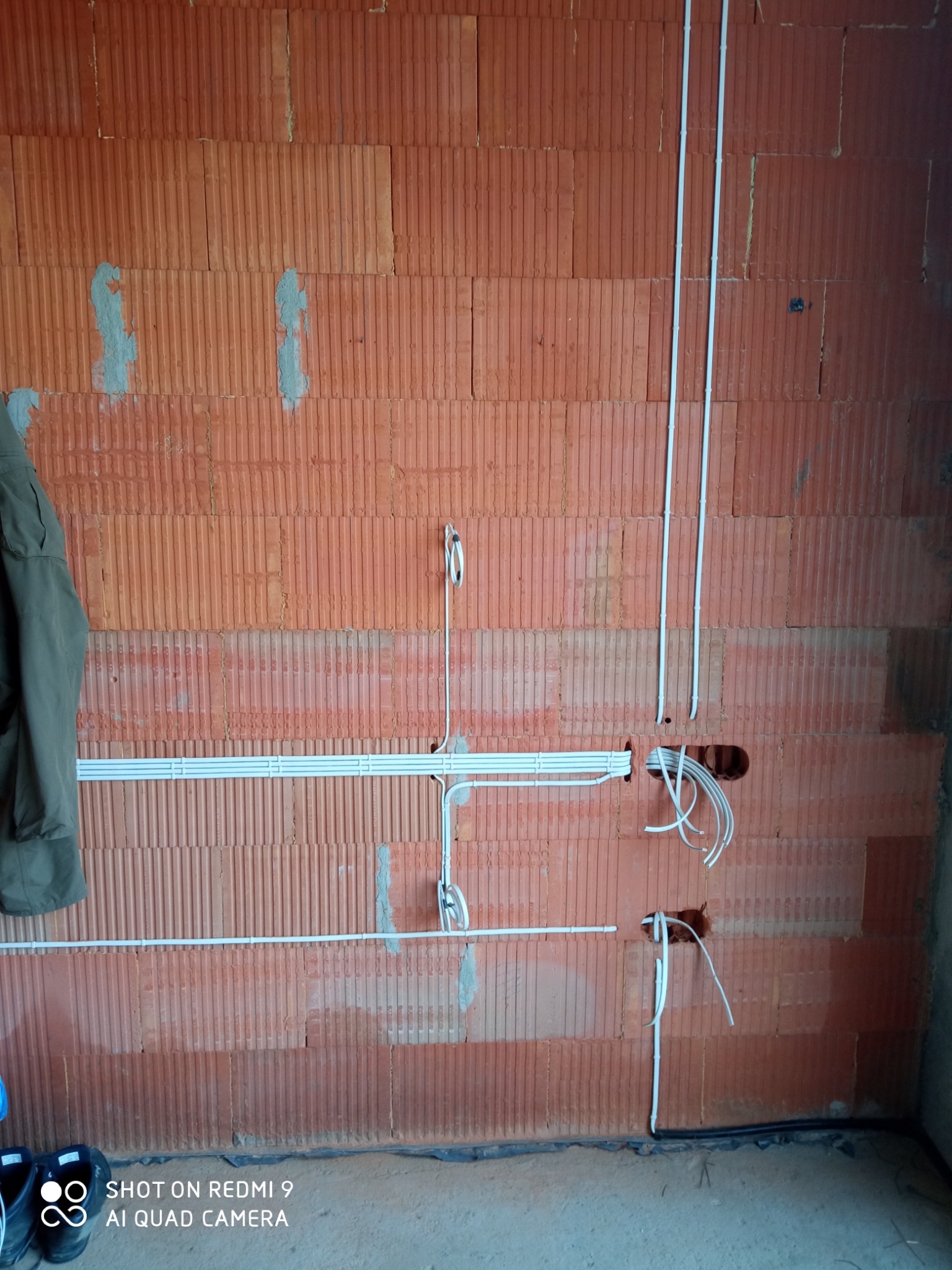 Ściana z cegły dziurawki z widocznymi przewodami elektrycznymi w białych rurkach instalacyjnych, wychodzącymi z otworów, oraz fragment odzieży roboczej i butów w dolnym rogu.