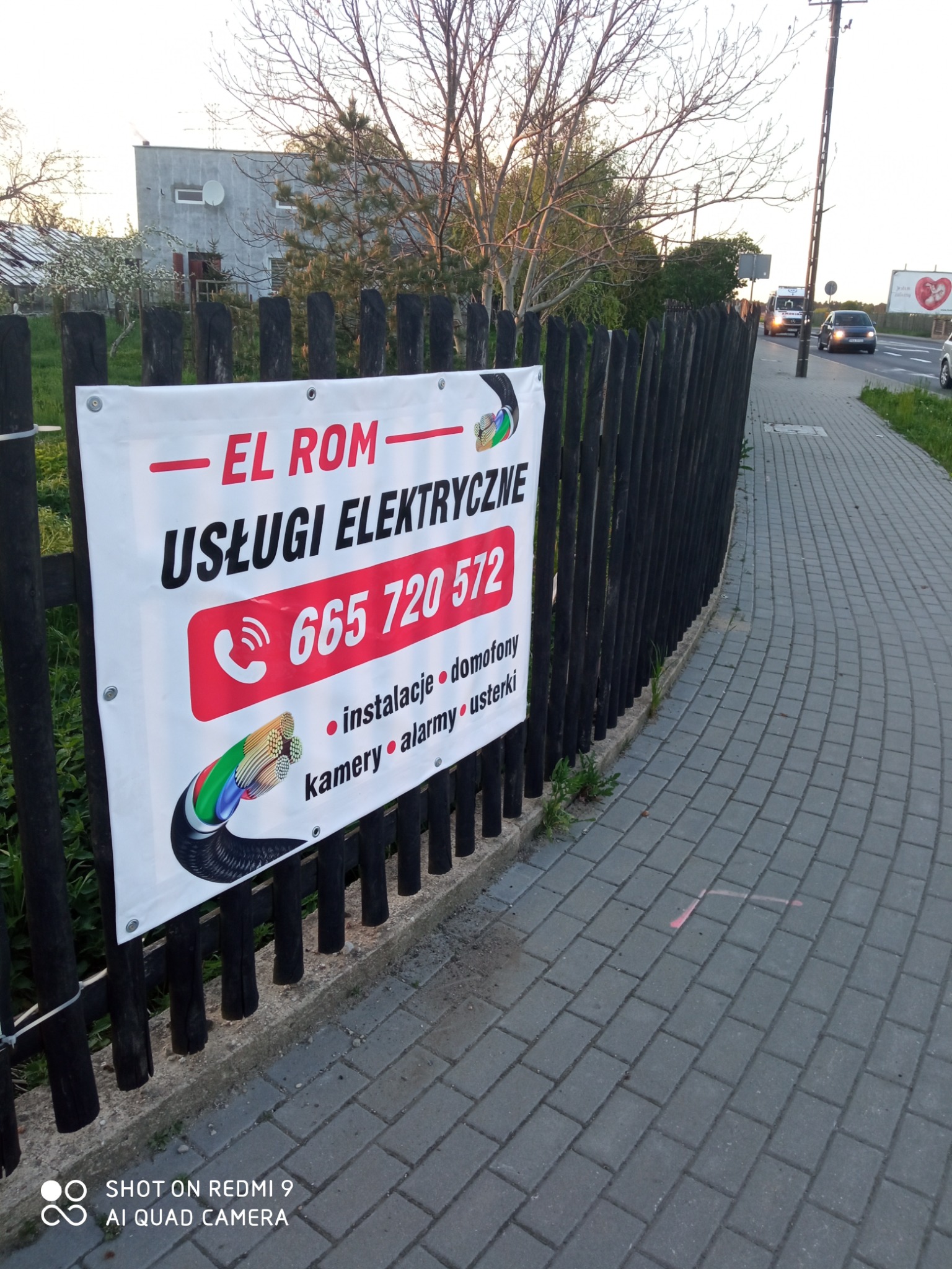 Baner reklamowy firmy 'EL ROM' oferującej usługi elektryczne, zawieszony na drewnianym płocie przy chodniku. Na banerze widnieje numer telefonu i lista usług: instalacje, domofony, kamery, alarmy...