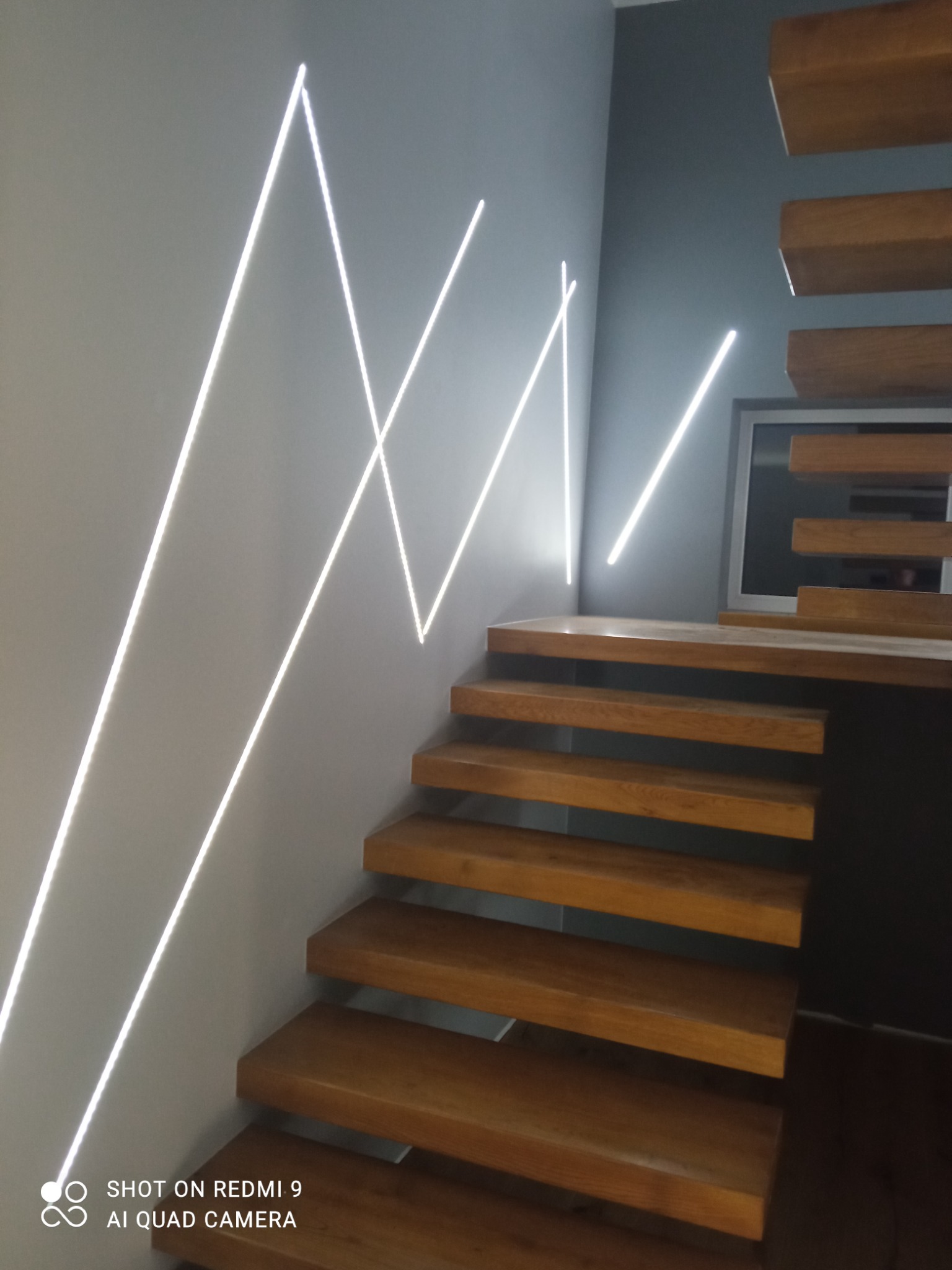 Nowoczesne schody drewniane z podświetleniem LED na ścianie w kształcie zygzaków, minimalistyczny design.