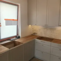 Wnętrze kuchni w trakcie montażu: szare szafki z drewnianym blatem, widoczne niedoskonałości ściany, okno z folią ochronną i planami, oświetlenie podszafkowe.