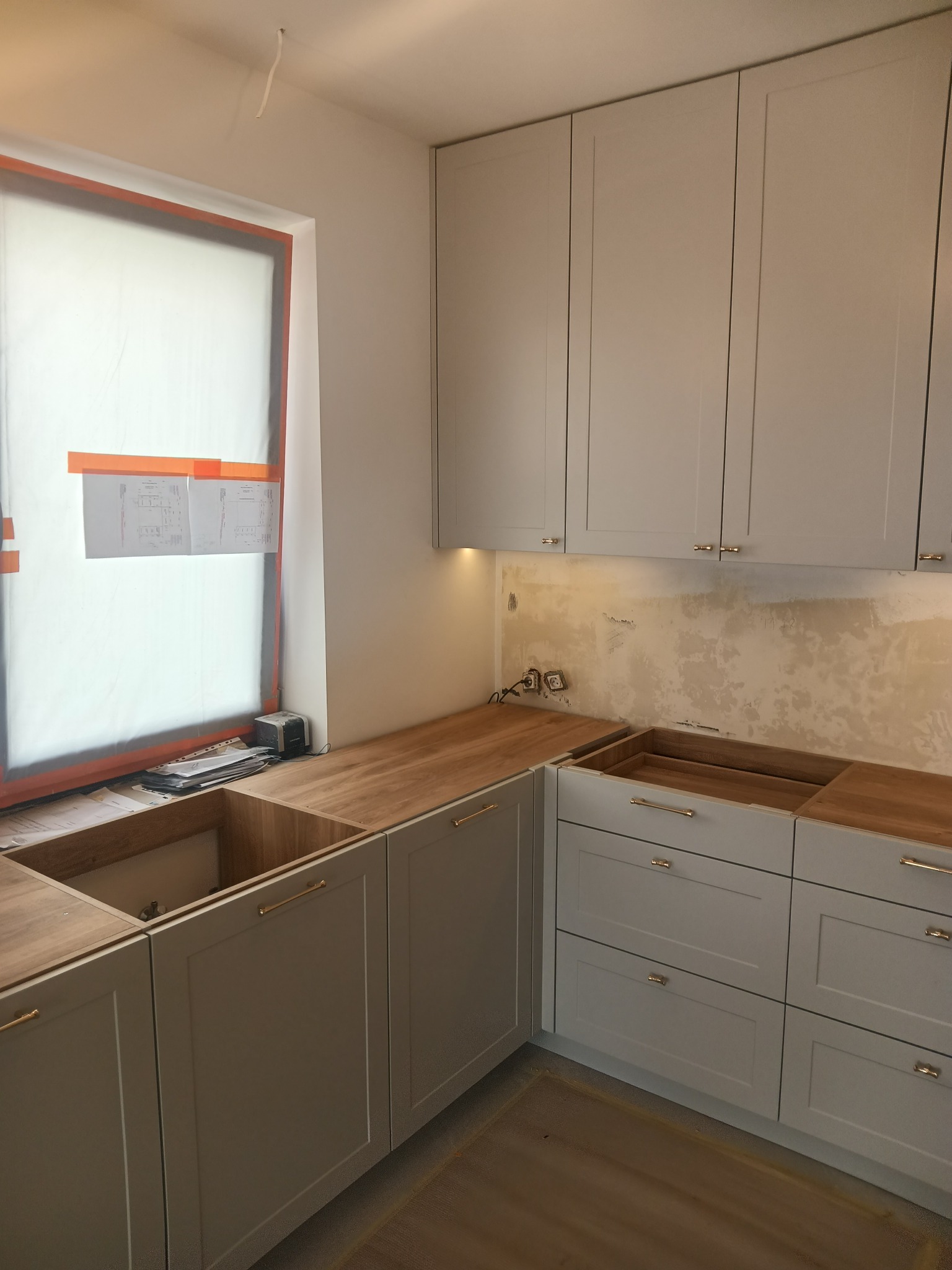 Wnętrze kuchni w trakcie montażu: szare szafki z drewnianym blatem, widoczne niedoskonałości ściany, okno z folią ochronną i planami, oświetlenie podszafkowe.