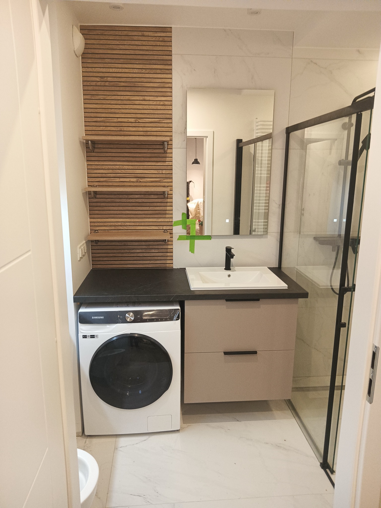 Nowoczesna łazienka z drewnianą ścianą, umywalką z szafką w kolorze beżowym, pralką Samsung i kabiną prysznicową z czarnymi ramami.