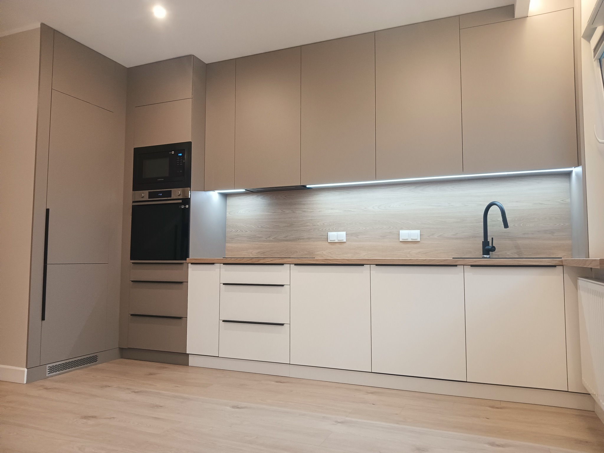 Nowoczesna kuchnia z beżowymi szafkami górnymi i białymi dolnymi, drewnianym blatem i czarną armaturą, oświetlenie LED pod szafkami.