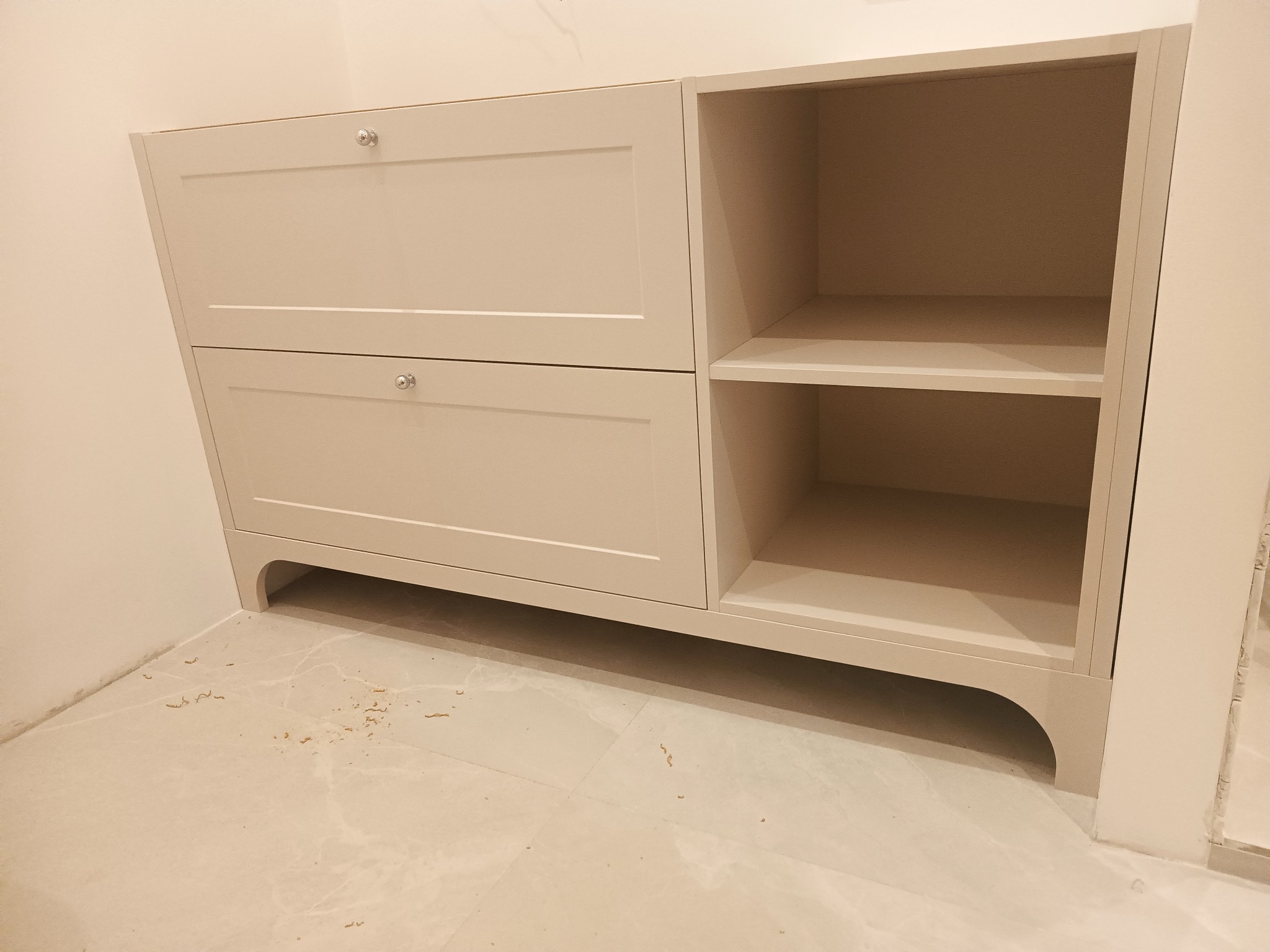 Jasny, beżowy mebel z dwiema szafkami z uchwytami i otwartą półką, widoczne ślady po montażu na podłodze.