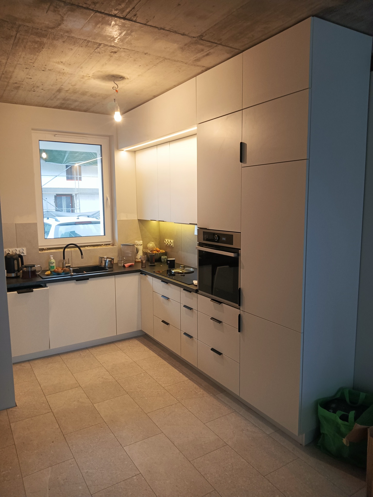 Nowoczesna kuchnia z białymi, matowymi szafkami i czarnymi uchwytami, oświetleniem LED pod szafkami wiszącymi, czarnym blatem i surowym betonowym sufitem z wiszącą żarówką.