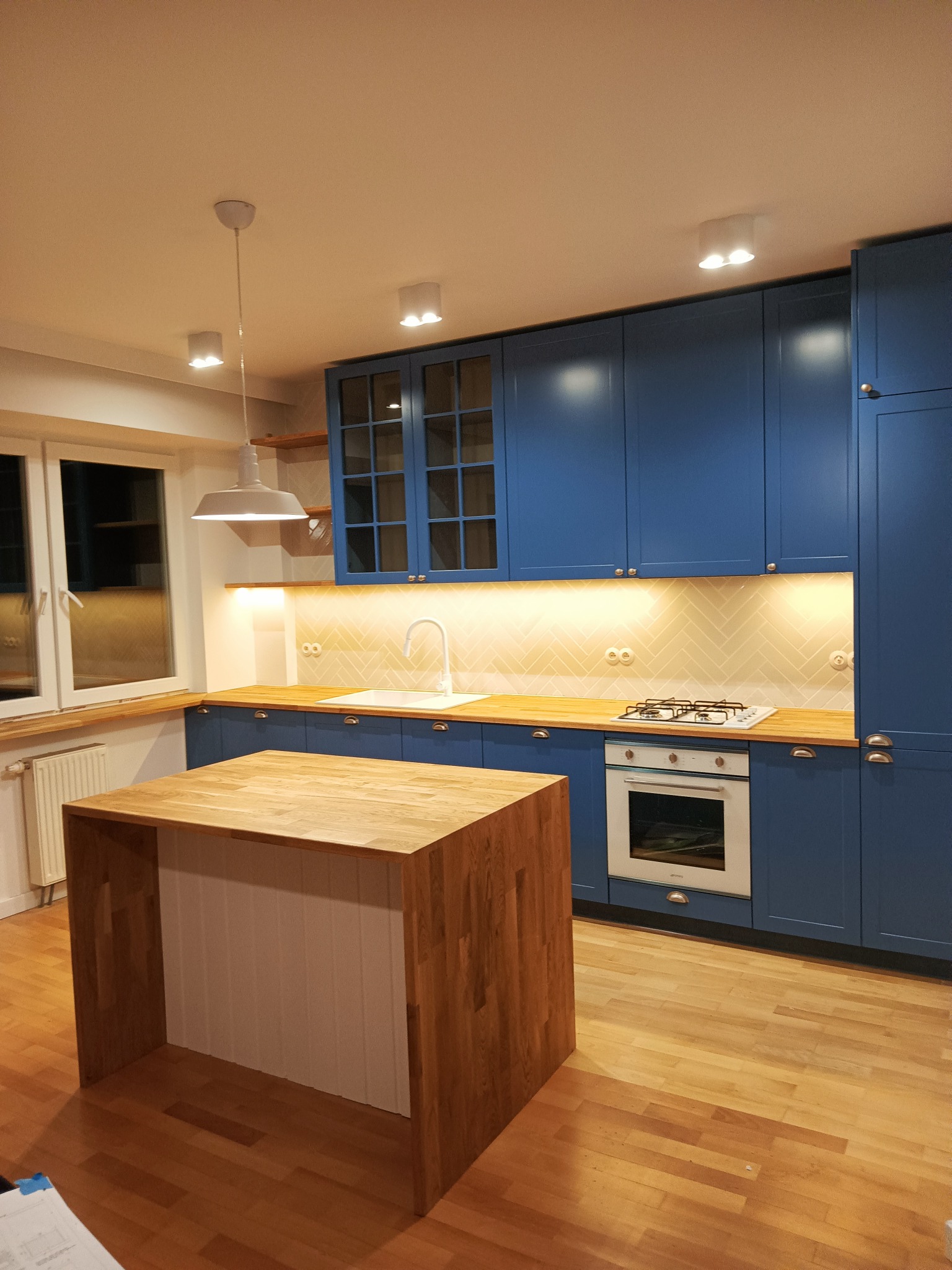 Niebieska zabudowa kuchenna z drewnianym blatem i wyspą, oświetlenie podszafkowe, białe płytki ułożone w jodełkę na ścianie nad blatem, nowoczesna lampa wisząca.