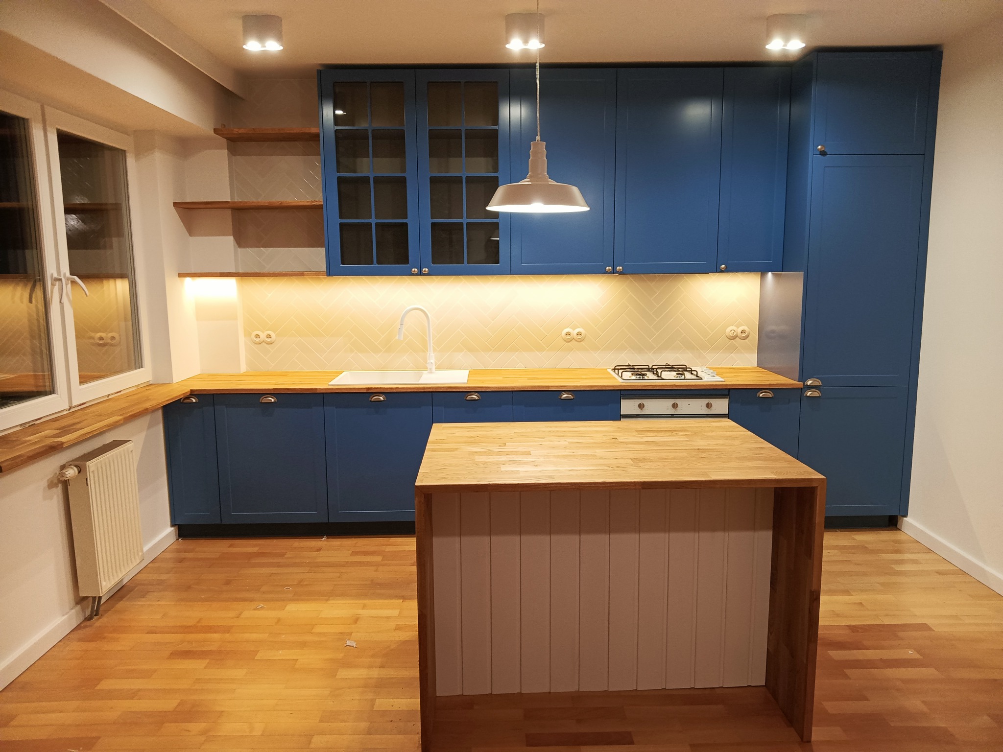Niebieska zabudowa kuchenna z drewnianym blatem, wyspą i oświetleniem punktowym oraz wiszącą lampą w stylu industrialnym, na tle parkietu.