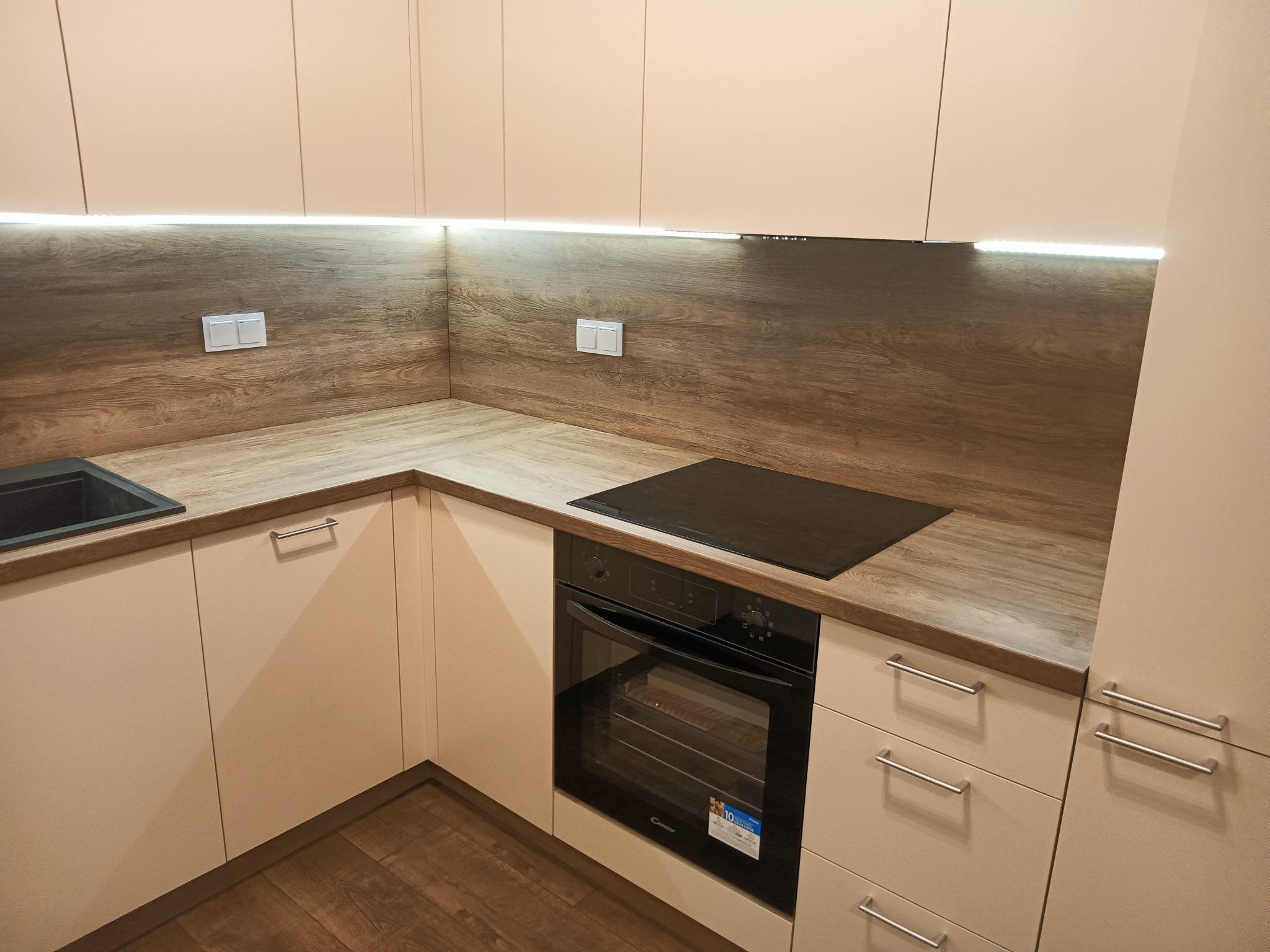 Jasna, nowoczesna zabudowa kuchenna z drewnopodobnym blatem, oświetleniem LED podszafkowym i czarną płytą indukcyjną.