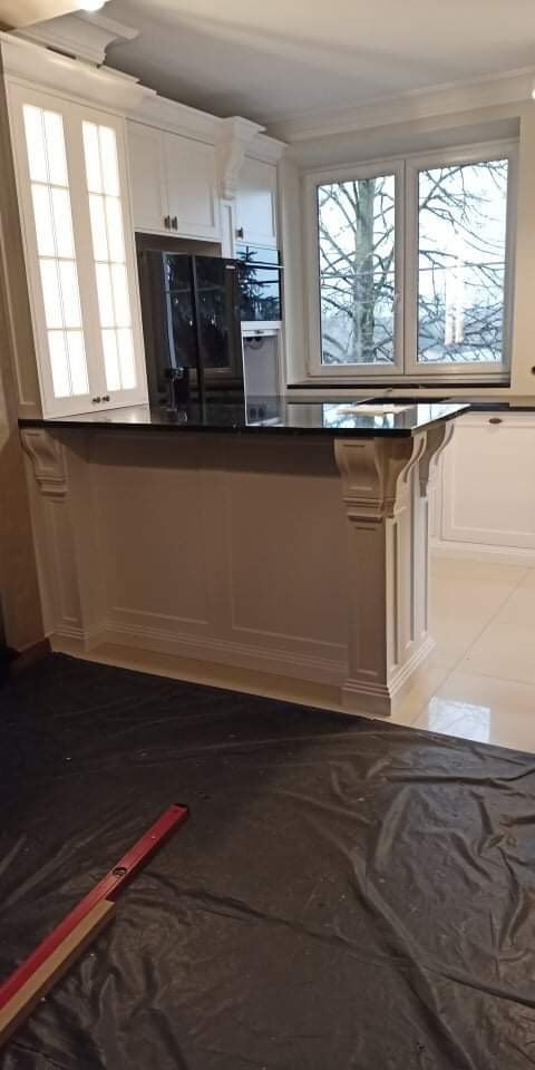 Wnętrze kuchni w trakcie montażu, widoczna biała wyspa z czarnym blatem i dekoracyjnymi wspornikami, zabezpieczona podłoga z widoczną łatą murarską.