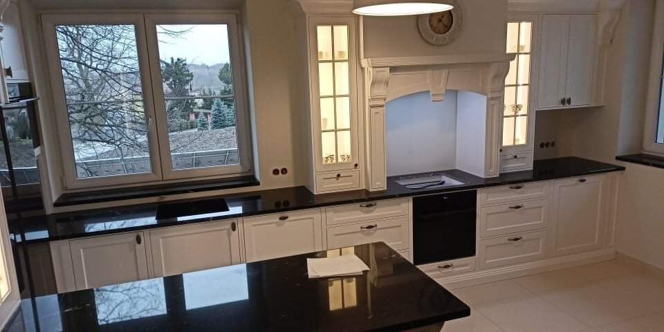 Klasyczna, biała zabudowa kuchenna z czarnymi blatami, wyspą i oknem z widokiem na drzewa bez liści, z subtelnym oświetleniem szafek.