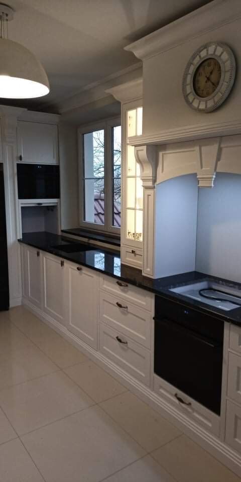 Klasyczna zabudowa kuchenna w odcieniach bieli z czarnym blatem, widoczny zegar na ścianie i okno z widokiem na zewnątrz.