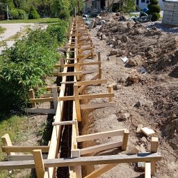 Michał Jurewicz - Przygotowanie do wylewki fundamentu pod ogrodzenie betonowe: wykopany rów z drewnianym szalunkiem i widocznym zbrojeniem stalowym, ziemia wokół, w tle dom i zieleń.