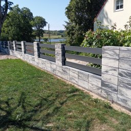 Michał Jurewicz - Nowoczesne ogrodzenie z szarej, wzorzystej kostki betonowej z poziomymi, grafitowymi przęsłami na tle zielonego trawnika, domu i jeziora w oddali.