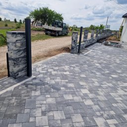 Michał Jurewicz - Nowoczesne ogrodzenie z betonowych bloczków w odcieniach szarości, z metalowymi słupkami, w trakcie montażu. Widoczna ułożona kostka brukowa i elementy krajobrazu.