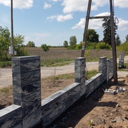 Michał Jurewicz - Fragment budowy ogrodzenia betonowego z szarej, wzorzystej kostki; widoczne słupki i przęsła w trakcie montażu, ziemia, słupy energetyczne w tle, słoneczny dzień.