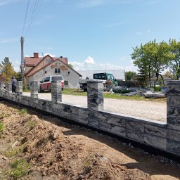 Michał Jurewicz - Wznoszenie ogrodzenia modułowego z szarego kamienia. Widoczny fragment konstrukcji z filarami, przygotowany teren i osoba w tle. W oddali dom i pojazdy.