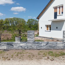 Michał Jurewicz - Niski murek z szarej, wzorzystej kostki betonowej, tworzący ogrodzenie posesji z białym domem w tle. Widoczny bęben z kablem elektrycznym.