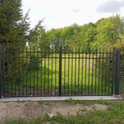 Nowoczesna, czarna brama metalowa z pionowymi szczebelkami, w tle zieleń drzew i trawy. Brama zamontowana na betonowej podmurówce.