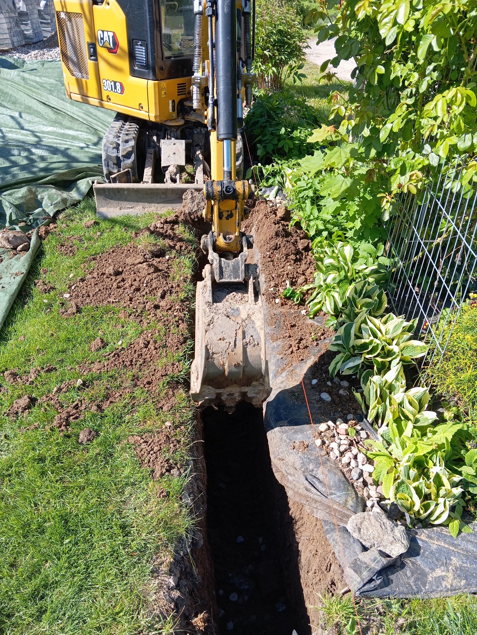 Żółta koparka CAT 301.8 wykopuje wąski rów w ziemi wzdłuż siatki ogrodzeniowej, widoczne trawnik, rabata z roślinami i folia ochronna.