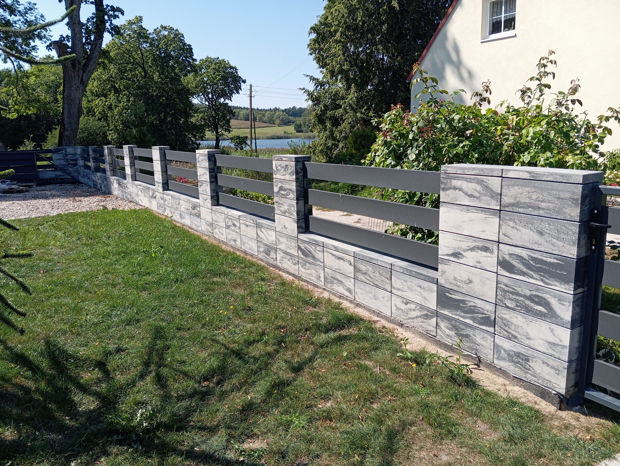 Nowoczesne ogrodzenie z szarej, wzorzystej kostki betonowej z poziomymi, grafitowymi przęsłami na tle zielonego trawnika, domu i jeziora w oddali.