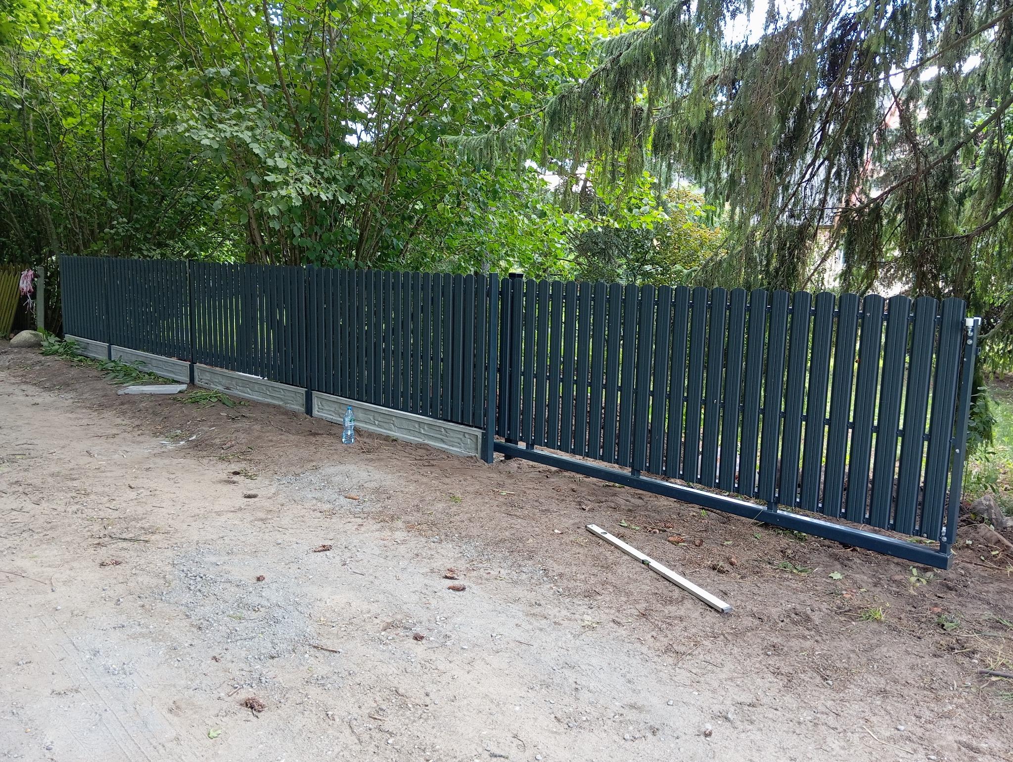 Nowoczesne, metalowe ogrodzenie panelowe w kolorze grafitowym z betonową podmurówką, widok z zewnątrz posesji, na ziemi leży poziomica, w tle zieleń drzew.