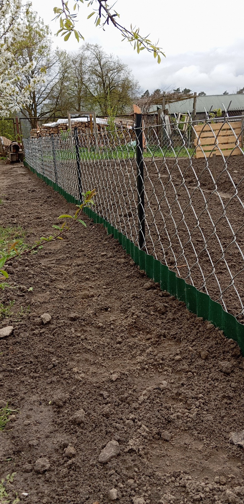 Długi fragment siatki ogrodzeniowej z zieloną, falistą osłoną dolną, rozciągnięty wzdłuż brązowej, świeżo przekopanej ziemi w ogrodzie.