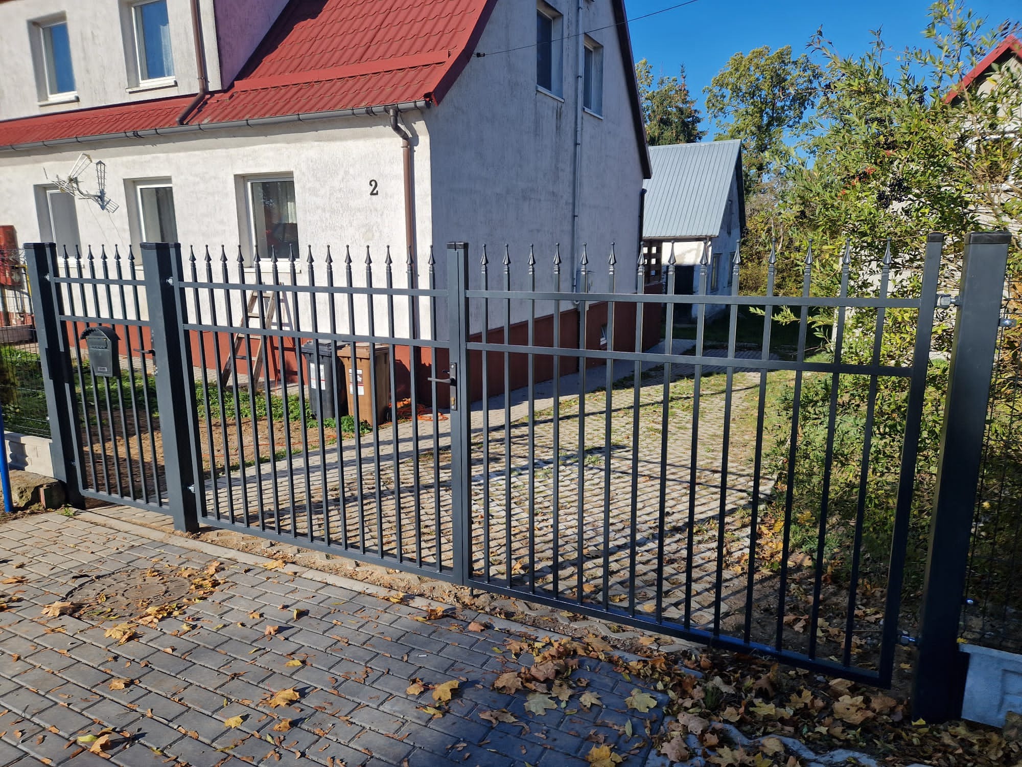 Czarna, metalowa brama wjazdowa z pionowymi prętami i ozdobnymi grotami, prowadząca na posesję z kostki brukowej, na której leżą opadłe liście.