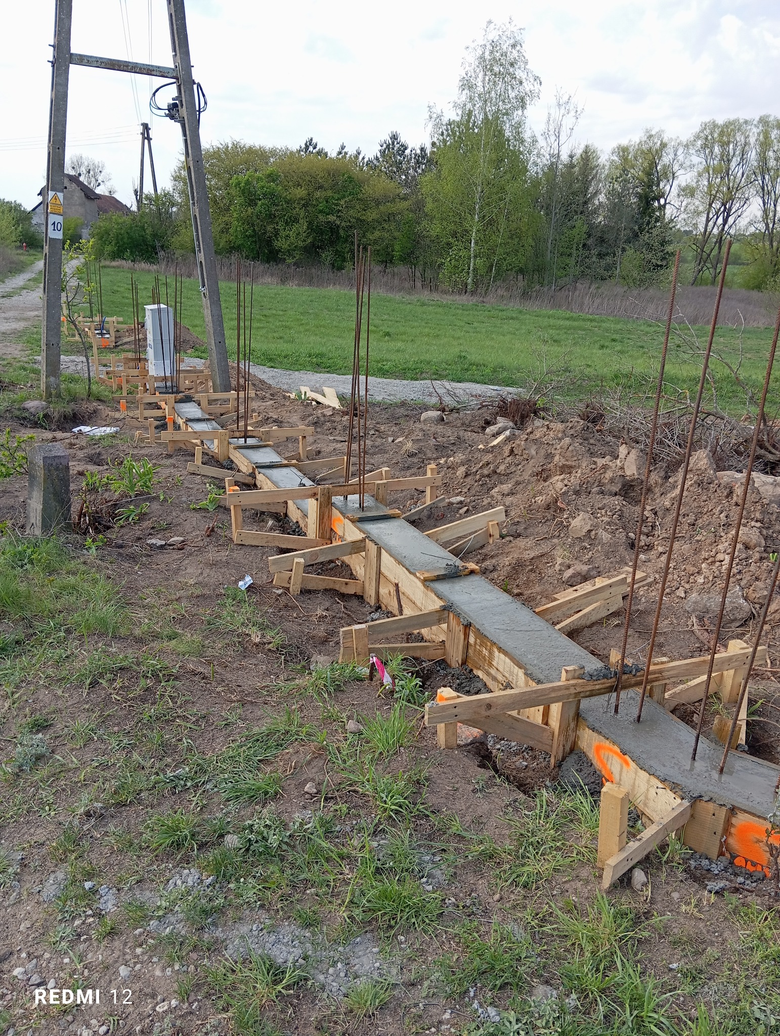 Wykop pod fundament ogrodzenia z drewnianym szalunkiem wypełnionym świeżym betonem i wystającymi prętami zbrojeniowymi, widok z poziomu gruntu, w tle słup energetyczny i zieleń.