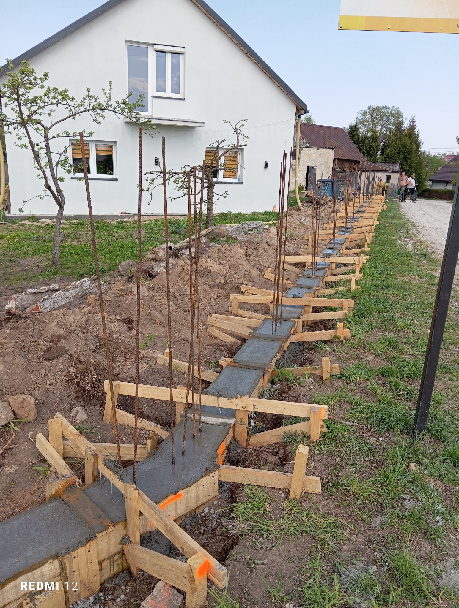 Wykop i szalunek pod fundament ogrodzenia z wystającymi prętami zbrojeniowymi, w tle dom jednorodzinny i zabudowania gospodarcze.