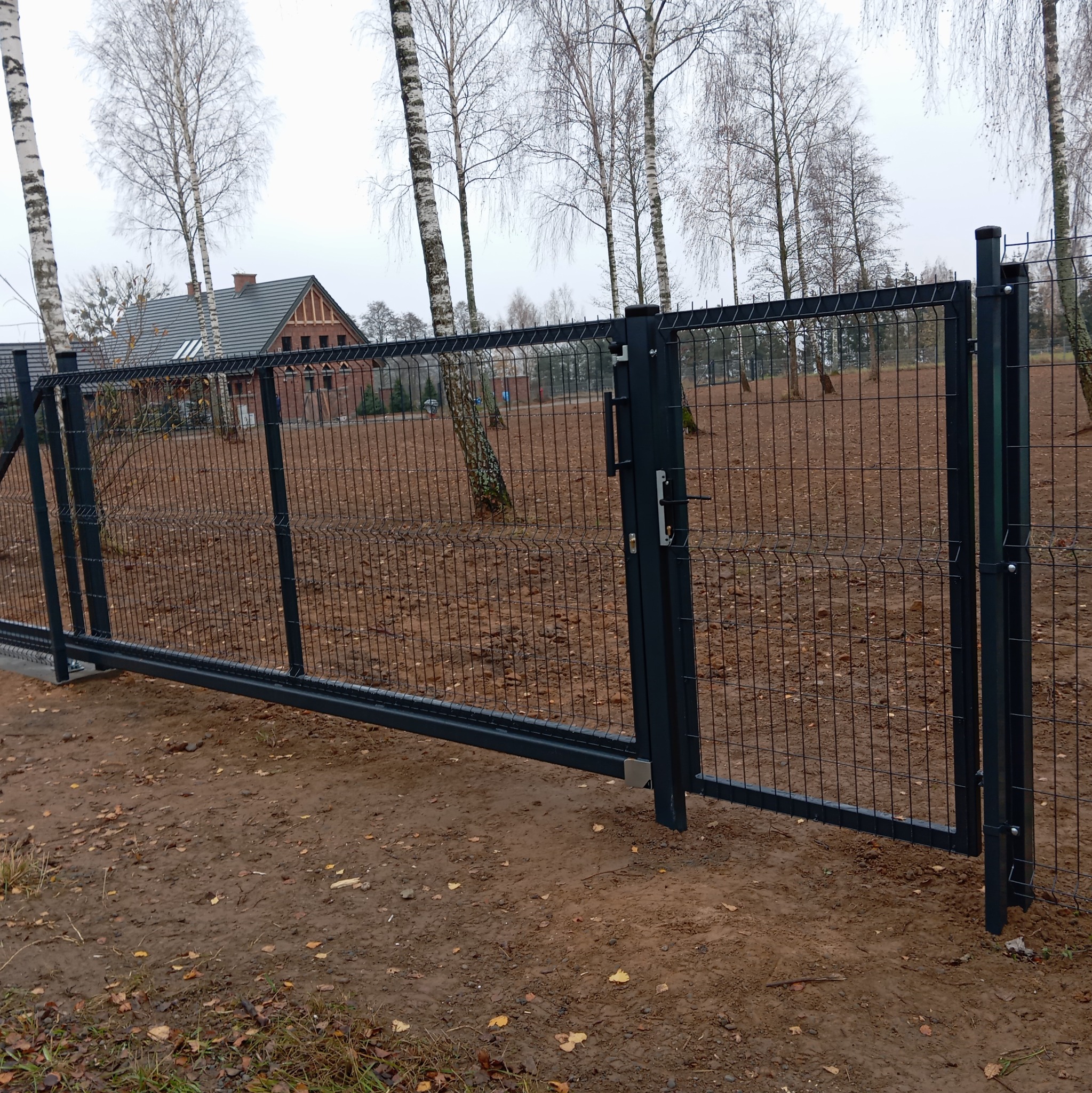 Przesuwna brama wjazdowa z paneli z widocznym zamkiem i uchwytem, na tle jesiennego krajobrazu z brązową ziemią i drzewami.