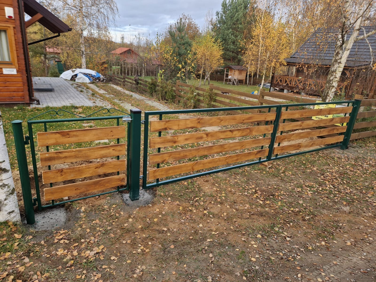 Nowoczesne ogrodzenie drewniane z zielonymi metalowymi słupkami i ramą, widoczne świeże betonowe fundamenty, jesienne otoczenie z domami w tle.