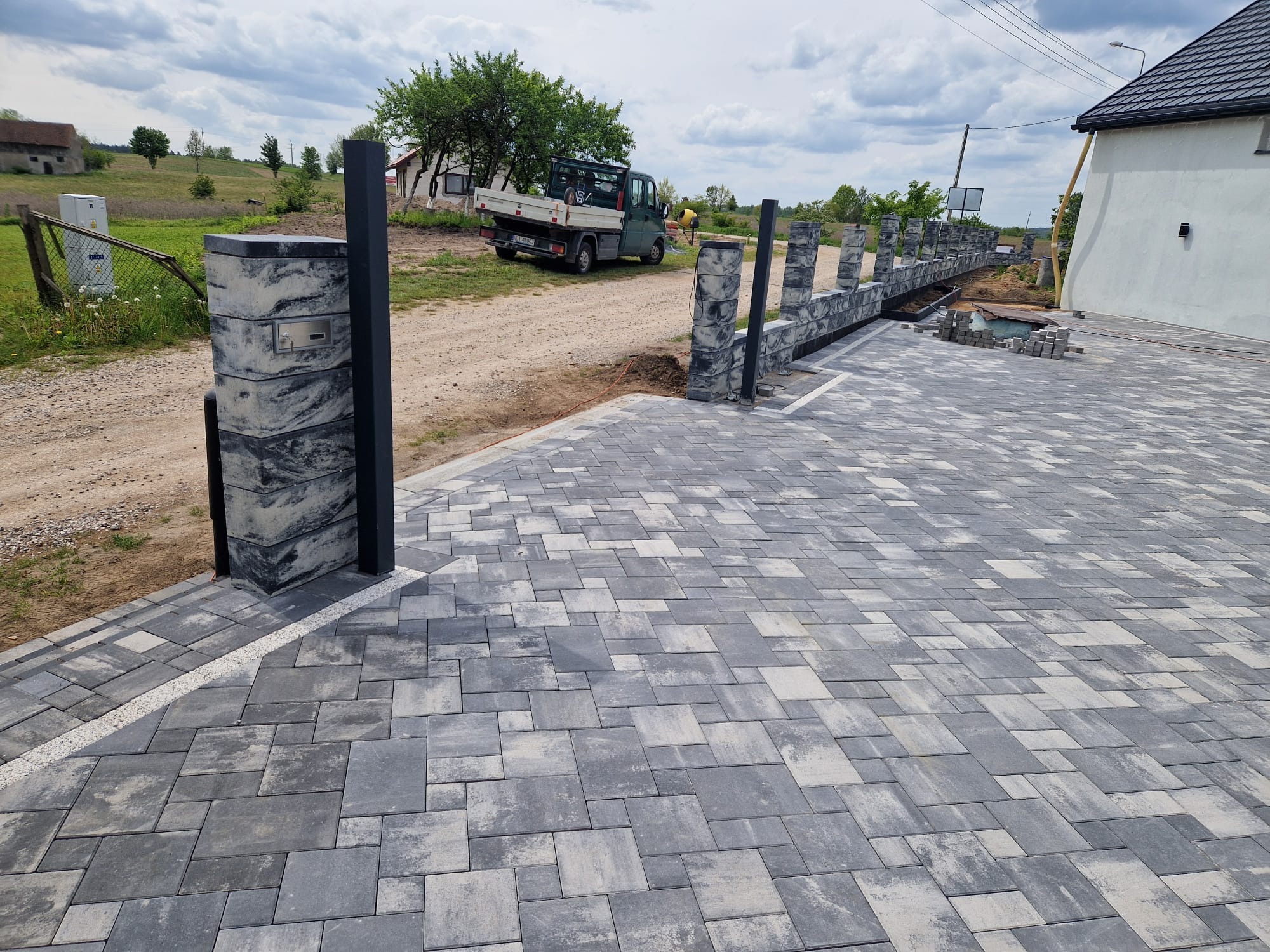 Nowoczesne ogrodzenie z betonowych bloczków w odcieniach szarości, z metalowymi słupkami, w trakcie montażu. Widoczna ułożona kostka brukowa i elementy krajobrazu.