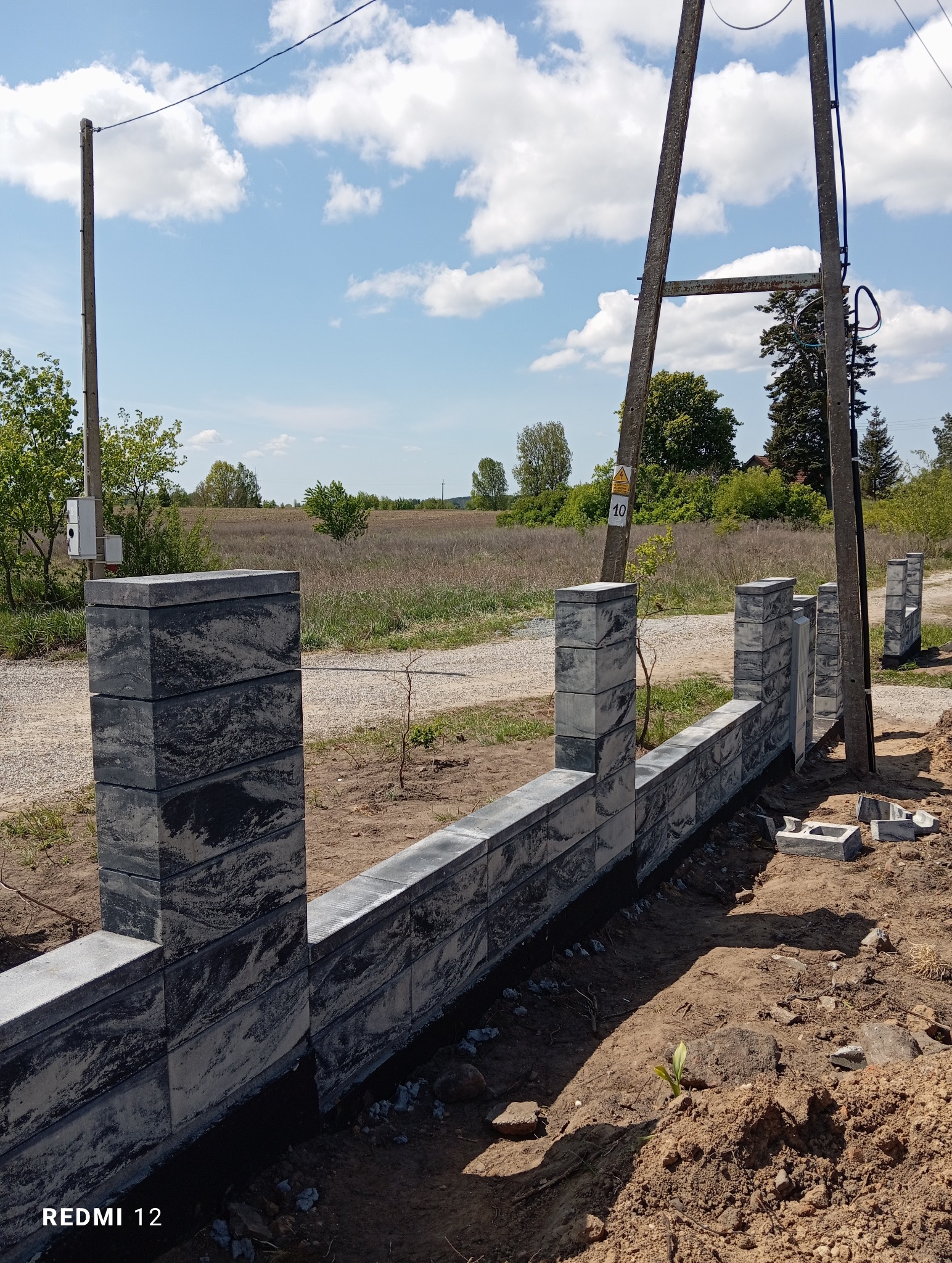 Fragment budowy ogrodzenia betonowego z szarej, wzorzystej kostki; widoczne słupki i przęsła w trakcie montażu, ziemia, słupy energetyczne w tle, słoneczny dzień.