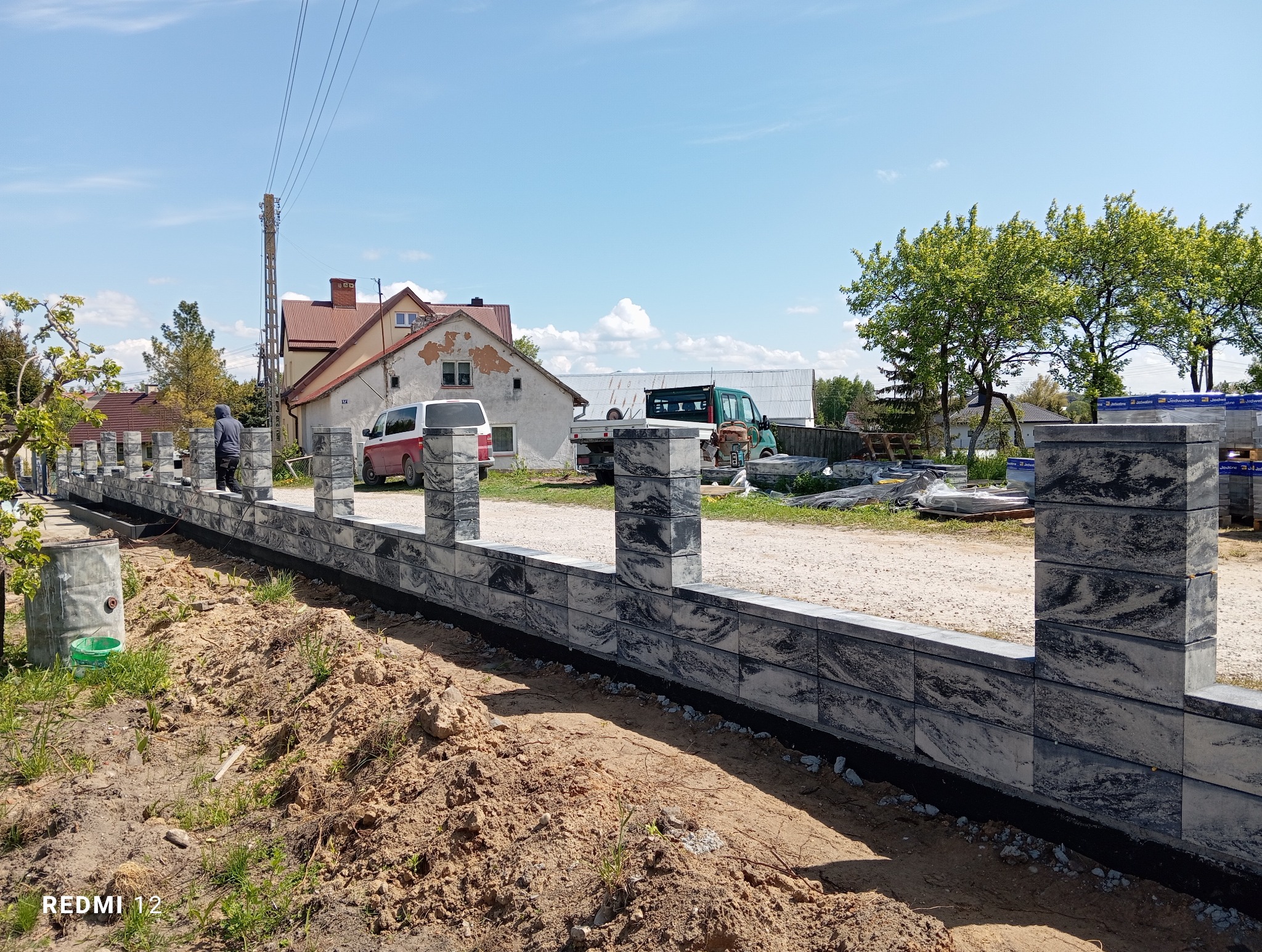 Wznoszenie ogrodzenia modułowego z szarego kamienia. Widoczny fragment konstrukcji z filarami, przygotowany teren i osoba w tle. W oddali dom i pojazdy.