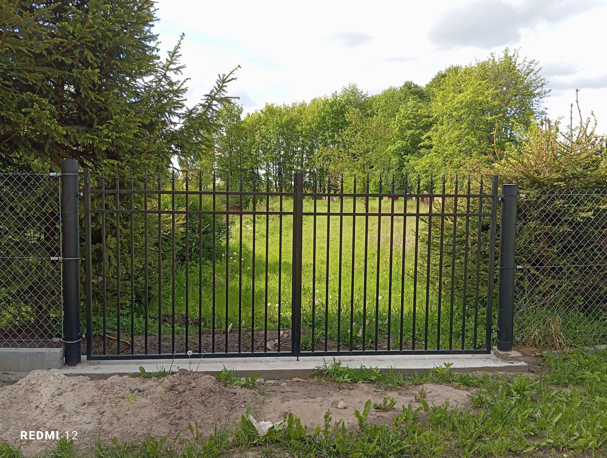 Nowoczesna, czarna brama metalowa z pionowymi szczebelkami, w tle zieleń drzew i trawy. Brama zamontowana na betonowej podmurówce.