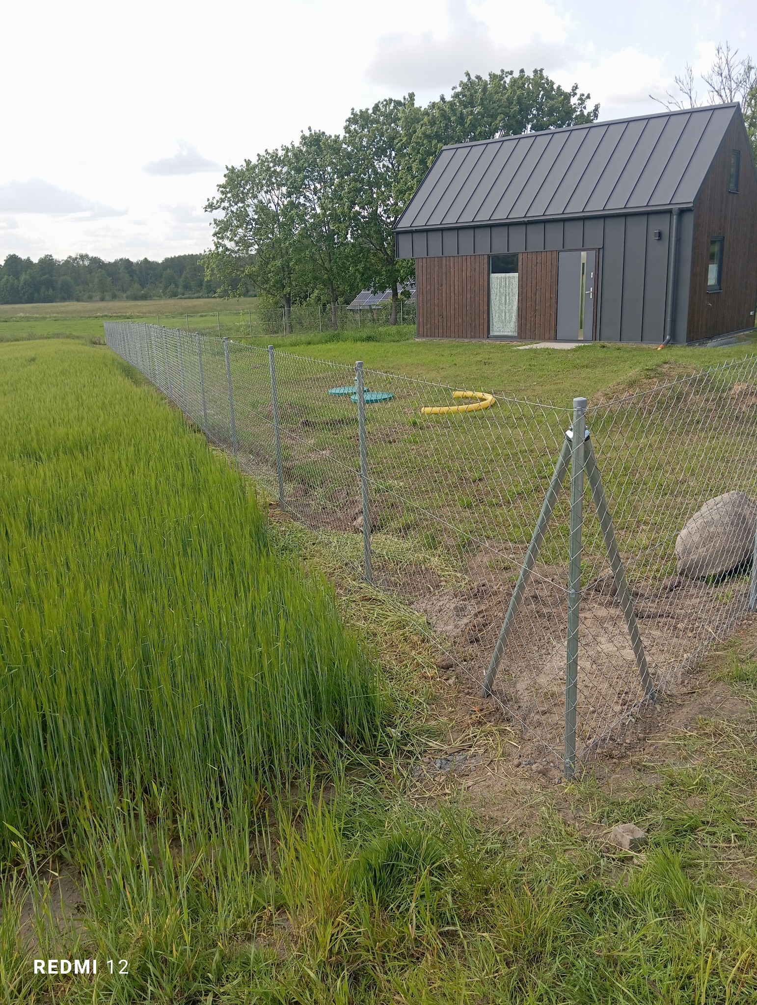 Widok na posesję z siatką ogrodzeniową i słupkami, za którą widać nowoczesny dom z ciemnym dachem i drewnianymi elementami elewacji, na tle zieleni i pochmurnego nieba.