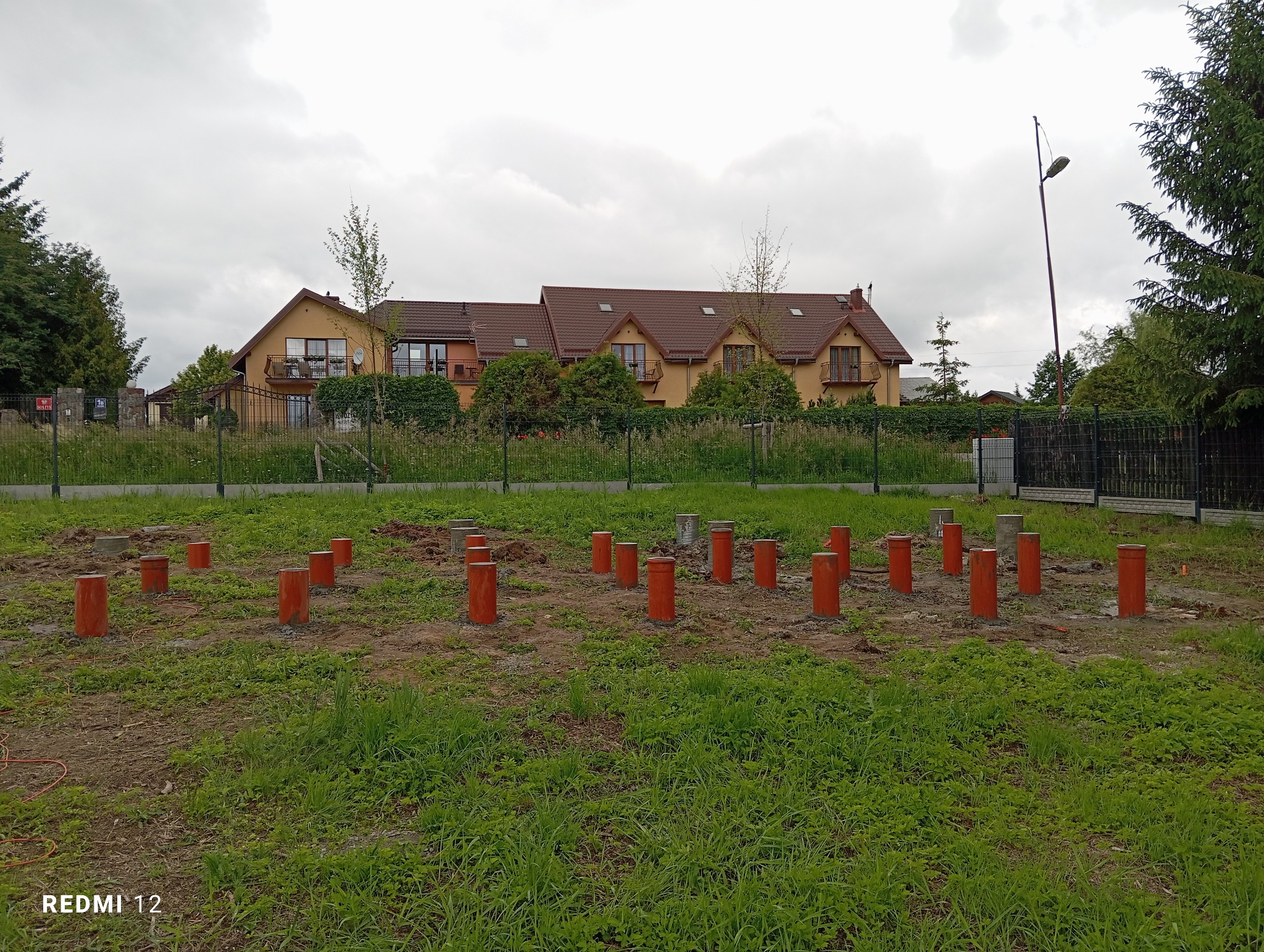 Przygotowanie terenu pod fundamenty ogrodzenia: pomarańczowe rury osadzone w ziemi na tle domu i istniejącego ogrodzenia z siatki.