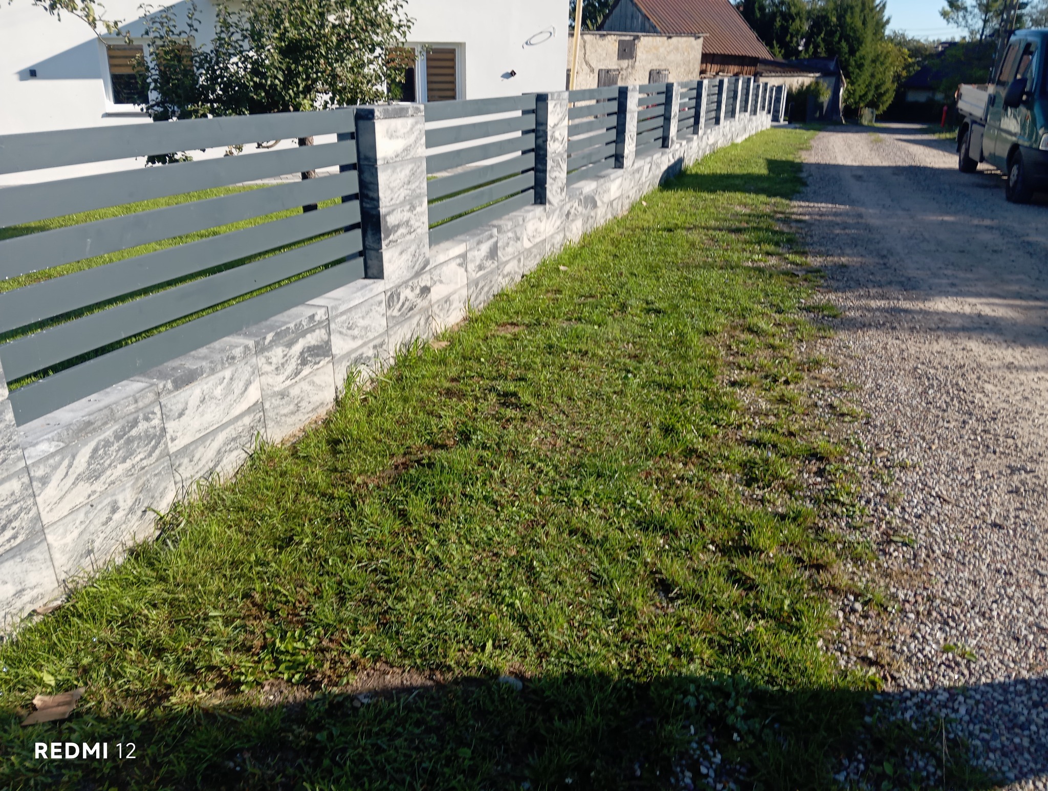 Nowoczesne ogrodzenie z poziomych paneli w kolorze grafitowym, osadzone na niskiej podmurówce z szarego kamienia, oddzielające posesję od żwirowej drogi.